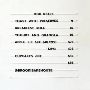 May include: A white sign with black text listing "BOX DEALS" including "TOAST WITH PRESERVES", "BREAKIEST ROLL", "YOGURT AND GRANOLA", "APPLE PIE 6PK", and "CUPCAKES 6PK". The sign also includes the text "@BROOKIBAKEHOUSE".