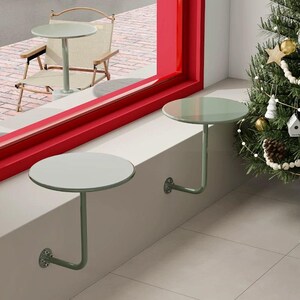May include: Two small, round, pale green tables mounted on a wall, with a matching table and chair visible outside a red-framed window. The tables have a minimalist design, with a single curved leg and a circular tabletop. A Christmas tree is in the background.