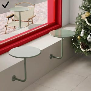 Puede incluir: Dos mesas redondas de color verde claro montadas en la pared con una sola pata curvada. Las mesas están fijadas a una pared blanca con un soporte metálico. Una tercera mesa es visible a través de un marco de ventana rojo, junto con una silla. Un árbol de Navidad está en el fondo.