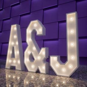 May include: White marquee letters A, &, and J with warm white lights. The letters are illuminated and stand on a reflective surface in front of a textured purple wall. The letters are a decorative element.