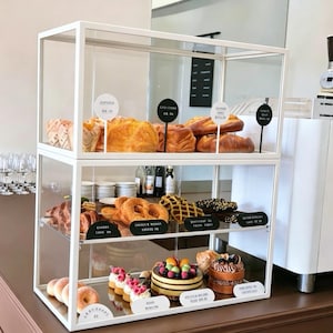 Könnte beinhalten: Eine zweistöckige Vitrine aus weißem Metall und Glas, gefüllt mit verschiedenen Backwaren wie Gebäck, Krapfen und Kuchen. Jeder Artikel ist mit kleinen runden oder rechteckigen Schildern versehen. Die Struktur hat klare Glasscheiben.