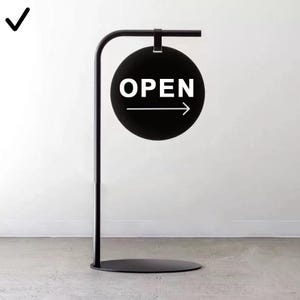 Peut inclure: Un panneau noir en métal avec un design circulaire. Le panneau affiche le mot "OPEN" en texte blanc avec une flèche pointant vers la droite. Le panneau est fixé à un support en métal noir.