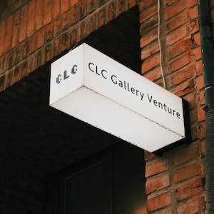 May include: A rectangular, illuminated sign with the text "CLC Gallery Venture" hangs from a brick wall. The sign is white with black lettering and the CLC logo in the upper left corner. The brick wall is a reddish-brown color.
