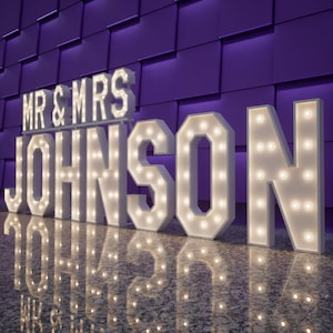 以下が含まれることがあります： 白く照らされた大きなマーキー文字で「MR & MRS JOHNSON」と綴られています。文字は温白色の電球で照らされ、輝く効果を生み出しています。背景は質感のある紫色の壁です。