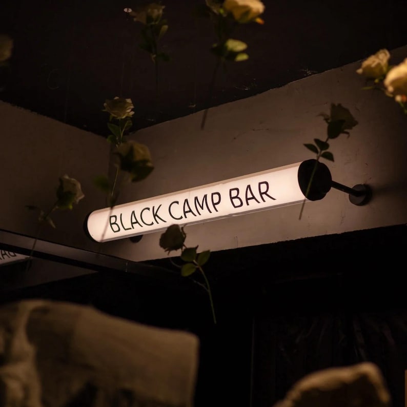 May include: A rectangular, illuminated sign with the words "BLACK CAMP BAR" in white lettering. The sign is mounted on a wall, and the background is dark. Artificial flowers hang from the ceiling.