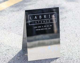 Puede incluir: Un letrero reflectante en ángulo con el texto "LABRIC STUDIO" en un marco rectangular blanco. Debajo, el letrero muestra el horario comercial y una dirección de correo electrónico. El letrero está sobre una superficie gris.