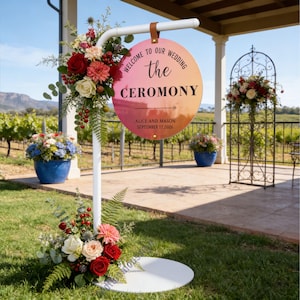 Può includere: Un cartello bianco per la cerimonia nuziale con un cartello circolare che recita "Welcome to our Wedding the Ceremony" con una freccia. Il cartello è decorato con fiori e verde. Il cartello include anche i nomi "Alice e Mason" e la data "17 settembre 2026."