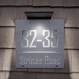 以下が含まれることがあります： 「32-35」の数字と「Strines Road」のストリート名が切り抜かれた、シルバーの金属板を使用したモダンな照明付き住所サイン。サインは濃いグレーの背景に取り付けられ、黒い上部が付いています。