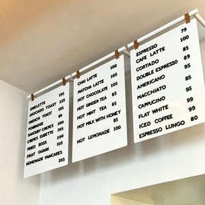 Op de afbeelding: Drie witte menuborden hangen aan een wit metalen frame met bruine leren riemen. De borden vermelden verschillende etenswaren en drankjes in zwarte tekst, waaronder espresso, omelet en chai latte.