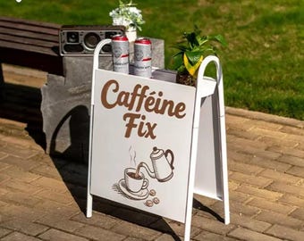 Puede incluir: Un letrero blanco en forma de A con las palabras "Caffeine Fix" en letras marrones. El letrero presenta una ilustración de una taza de café y una tetera. Dos latas de cerveza y una pequeña planta se encuentran en la parte superior.