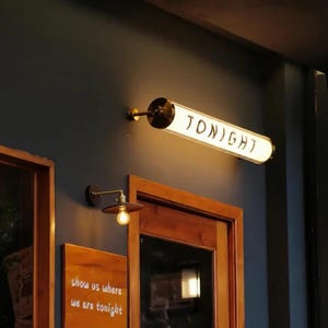 May include: A wall-mounted sign with the word "TONIGHT" in black lettering. The sign is illuminated and has a cylindrical shape with brass-colored hardware. Below, a small, round light fixture with a single bulb is mounted on the wall.