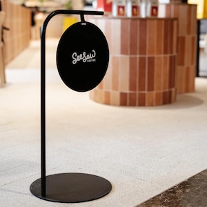 May include: A black metal sign with the text "Seesaw Coffee" in white script. The sign is oval-shaped and hangs from a curved metal pole. The pole is attached to a round base, and the sign is likely used for advertising.