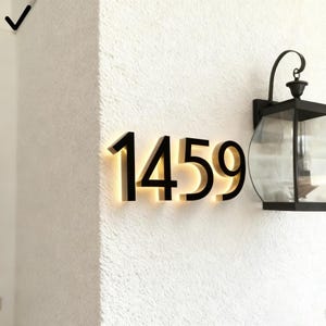 May include: Black illuminated house numbers 1459 mounted on a textured white wall. The numbers are backlit with a warm glow. A black outdoor lantern is mounted to the right of the numbers.