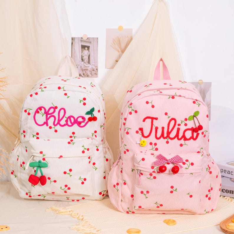May include: Two corduroy backpacks, one white and one pink, are decorated with a cherry pattern. The white backpack has the name "Chloe" embroidered in pink, while the pink backpack has "Julia" embroidered in red. Both have a front pocket and a top handle.