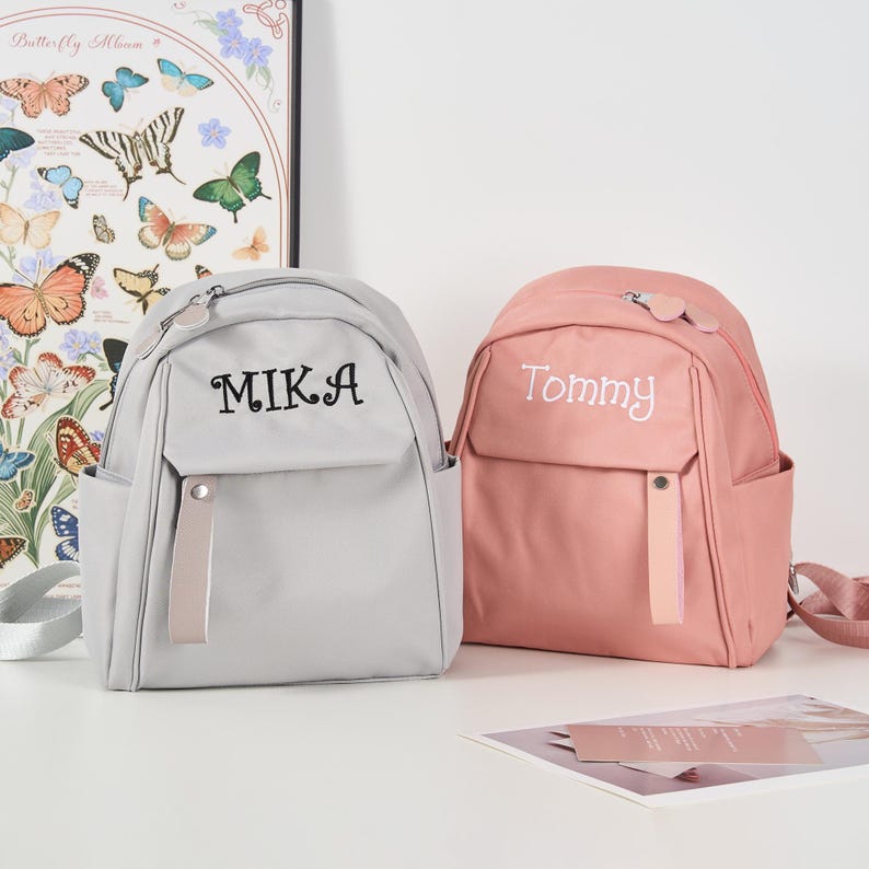Puede incluir: Dos mochilas personalizadas. Una es de color gris claro con el nombre "MIKA" bordado en negro. La otra es rosa con el nombre "Tommy" bordado en blanco. Ambas tienen un bolsillo frontal, una cremallera superior y una correa del mismo color.