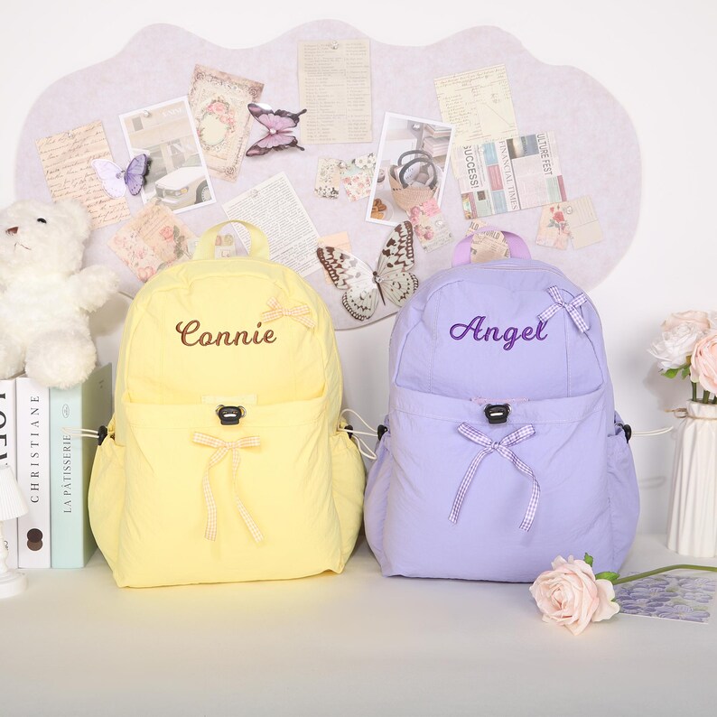 May include: Two nylon backpacks, one yellow and one lavender, are displayed. The yellow backpack has the name "Connie" embroidered on it, while the lavender backpack has "Angel" embroidered. Both have bow accents and side pockets. The background includes decorative butterflies and stationery.