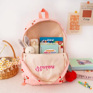 May include: Pink backpack with a cherry pattern, open to reveal books, a water bottle, and a stuffed bunny. The backpack has a red pom-pom and the word "Bonjour" embroidered on the front.