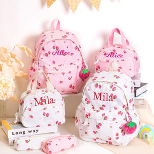 May include: Pink and white backpacks, lunch bags, and pencil cases with a strawberry pattern. The name "Alice" is embroidered on some bags, and "Mila" on others. Each bag has a strawberry charm.