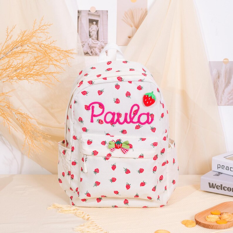 May include: White backpack with a strawberry pattern and the name "Paula" embroidered in pink. The backpack has a front pocket, side pockets, and a green strawberry patch. A bow with red and green accents is also on the front.