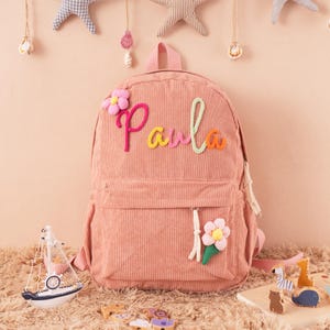 May include: Pink corduroy backpack with the name "Paula" embroidered in colorful thread. The backpack features a front pocket, a top handle, and a flower applique. Decorative elements include hanging starfish and a toy sailboat.