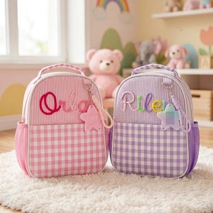May include: Two backpacks with top handles and side mesh pockets. The left backpack is pink with vertical stripes and a gingham bottom, featuring the name Orla. The right backpack is purple with vertical stripes and a gingham bottom, featuring the name Riley.