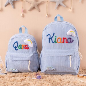 May include: Two light blue corduroy backpacks with rainbow-colored embroidered names and cloud patches. One backpack says "Qara" and the other says "Kiana". Both have a front pocket, a top handle, and a small turtle applique.