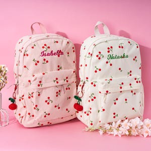 May include: Two white canvas backpacks with red cherry patterns. The backpack on the left has the name "Isabella" embroidered on it in pink. The backpack on the right has the name "Natasha" embroidered on it in green. Both backpacks have a small red cherry keychain attached.