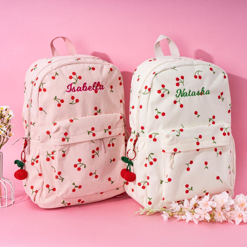 May include: Two white canvas backpacks with red cherry patterns. The backpack on the left has the name "Isabella" embroidered on it in pink. The backpack on the right has the name "Natasha" embroidered on it in green. Both backpacks have a small red cherry keychain attached.