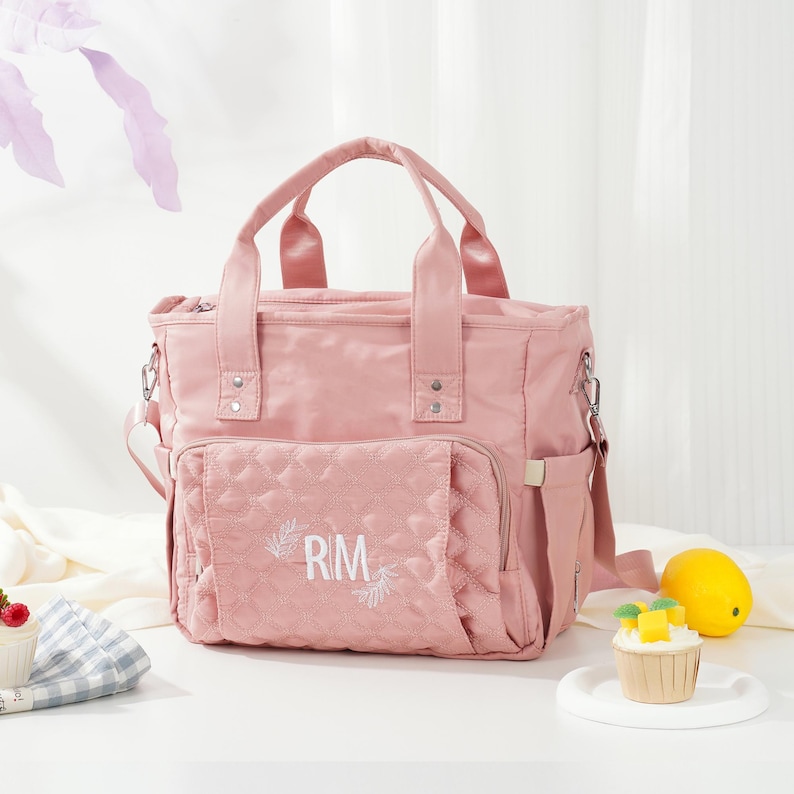 May include: A pink diaper bag with top handles and a shoulder strap. The front pocket features a quilted design with the embroidered initials "RM". The bag has multiple compartments and a side pocket. A lemon and a cupcake are in the background.