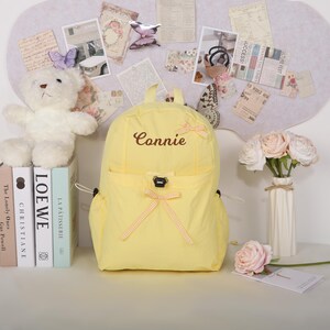 May include: A yellow backpack with the name "Connie" embroidered on the front. The backpack has a front pocket and is decorated with small bows. A white teddy bear, books, and flowers are also in the image.