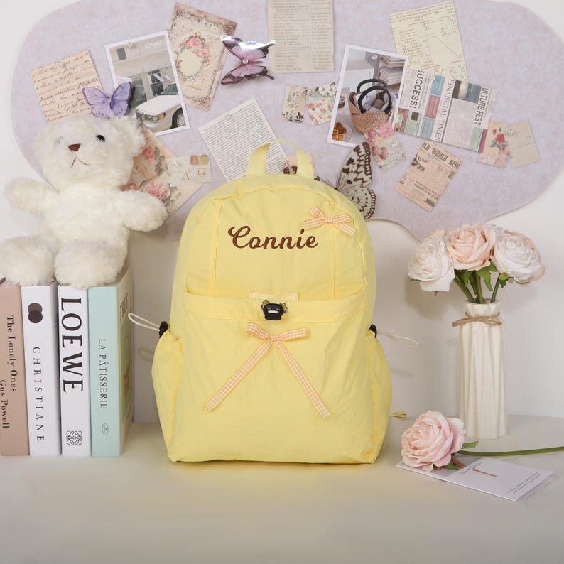 May include: A yellow backpack with the name "Connie" embroidered on the front. The backpack has a front pocket and is decorated with small bows. A white teddy bear, books, and flowers are also in the image.