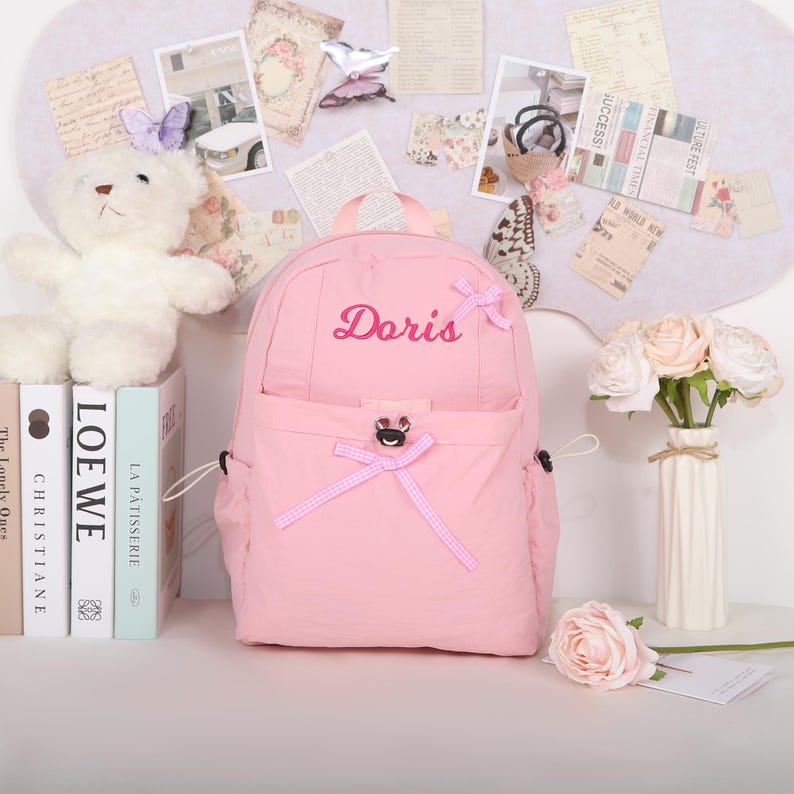May include: A pink backpack with the name "Doris" embroidered in cursive. The backpack has a front pocket with a bow and a top handle. A white teddy bear and books are to the left of the backpack. A vase of flowers is to the right.