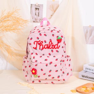 May include: Pink corduroy backpack with a strawberry pattern and the name "Thalida" embroidered in red. Features a front pocket, side pockets, and a red flower accent. The backpack has a top handle and adjustable straps.