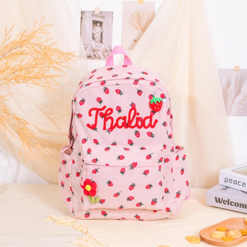 May include: Pink corduroy backpack with a strawberry pattern and the name "Thalida" embroidered in red. Features a front pocket, side pockets, and a red flower accent. The backpack has a top handle and adjustable straps.