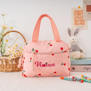 May include: Pink lunch bag with a cherry pattern and the name "Vivian" embroidered in cursive. The bag has a front pocket, a zipper closure, and a matching pink handle. A basket of flowers and a stuffed bunny are in the background.