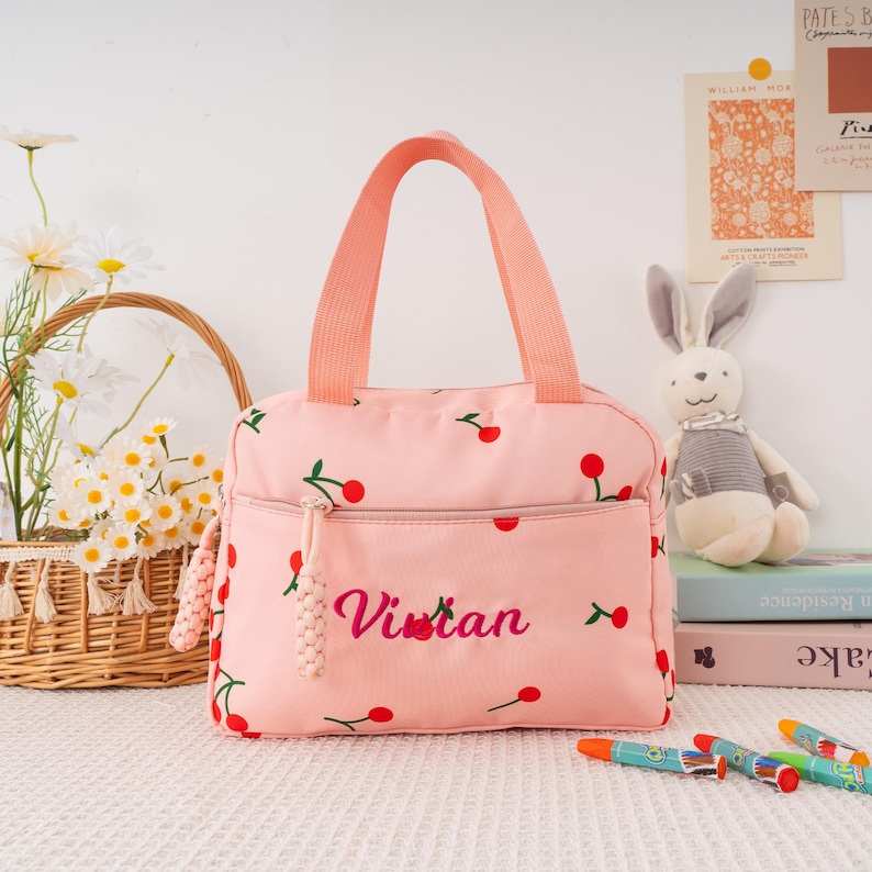 May include: Pink lunch bag with a cherry pattern and the name "Vivian" embroidered in cursive. The bag has a front pocket, a zipper closure, and a matching pink handle. A basket of flowers and a stuffed bunny are in the background.