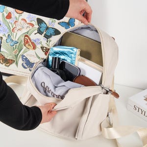 Puede incluir: Mochila beige con una correa de color claro y cremallera plateada. La mochila est&aacute; abierta, revelando varios objetos en su interior, incluyendo una bolsa azul, una cartera marr&oacute;n y una prenda gris. Una obra de arte con tem&aacute;tica de mariposas es visible en el fondo.