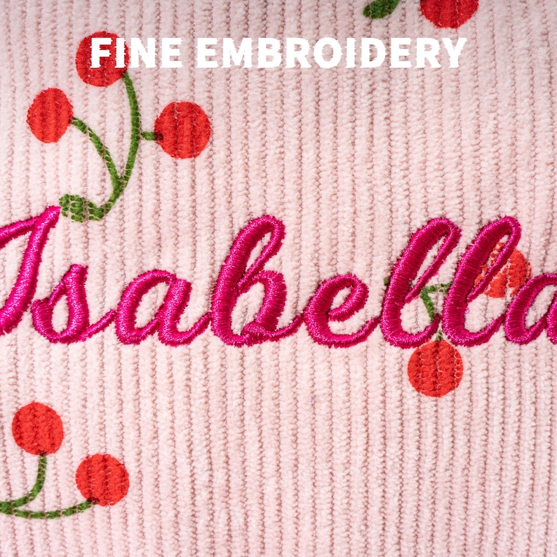 May include: Pink corduroy fabric with a cherry print and the name "Isabella" embroidered in pink thread. The text "FINE EMBROIDERY" is also visible at the top of the image.