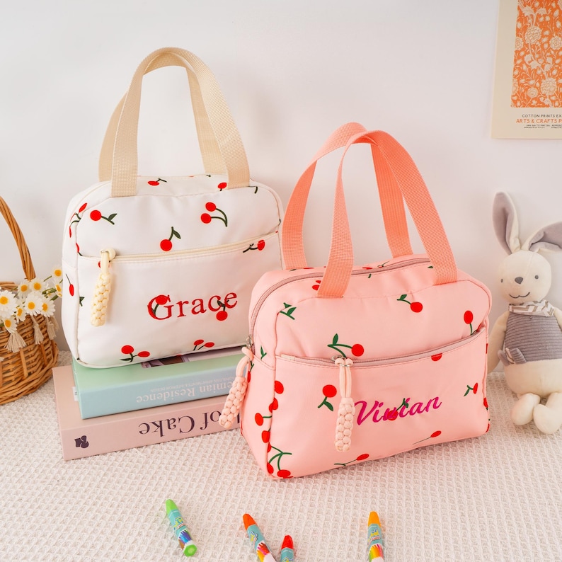 May include: Two lunch bags, one white and one pink, are displayed. Both bags feature a cherry pattern and a front zipper pocket. The white bag has "Grace" embroidered, while the pink bag has "Vivian". Each bag has a top handle and a decorative zipper pull.
