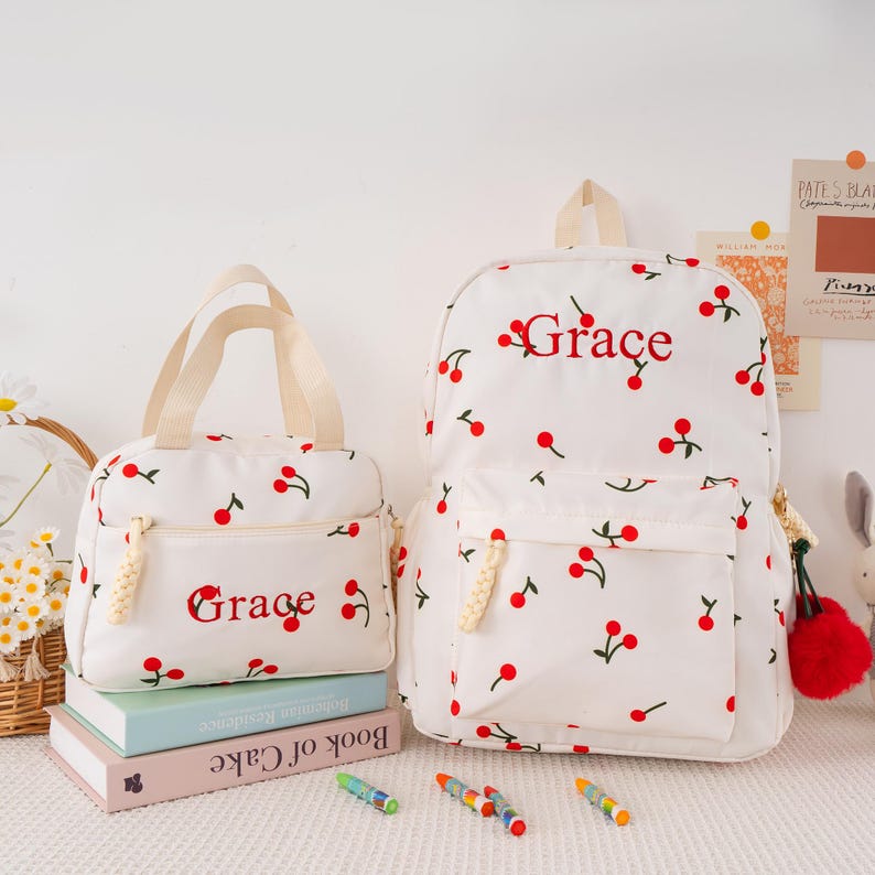 May include: White backpack and lunch bag set with a red cherry pattern. The word "Grace" is embroidered in red on both items. The lunch bag has a beige handle and zipper pulls. The backpack has a front pocket and a red pom-pom charm.
