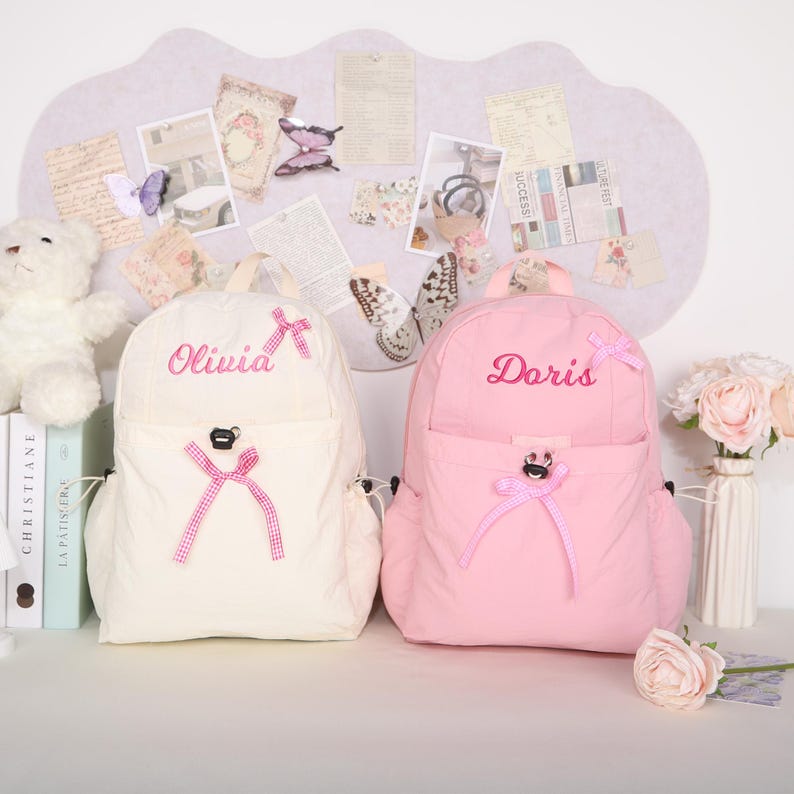 May include: Two backpacks, one cream and one pink, are displayed. The cream backpack has the name "Olivia" embroidered in pink, while the pink backpack has "Doris." Both have pink bows and bear-shaped accents.