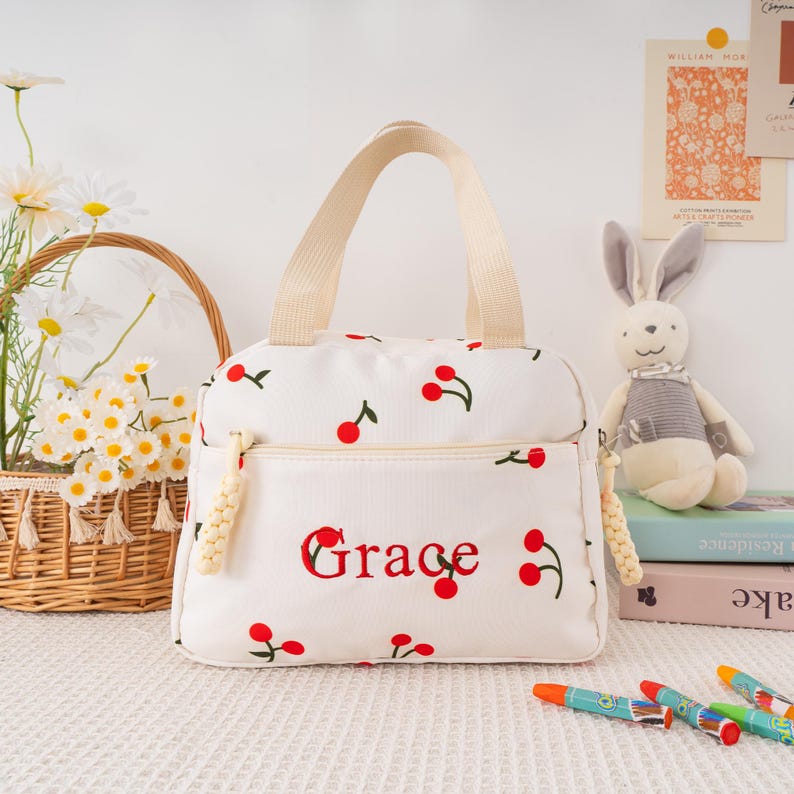 May include: White lunch bag with a cherry pattern and the name "Grace" embroidered in red. The bag has a beige handle and zipper, and a decorative tassel. A basket of flowers and a stuffed bunny are in the background.