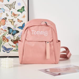 Puede incluir: Una mochila rosa empolvado con el nombre "Tommy" bordado en blanco. La mochila tiene un bolsillo frontal, bolsillos laterales y correas ajustables. Un estampado art&iacute;stico de mariposas es visible en el fondo.