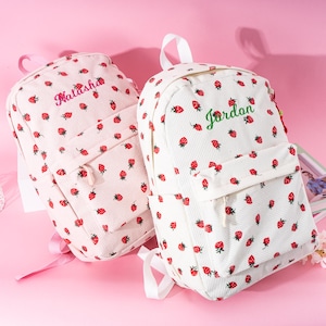 May include: Two corduroy backpacks, one light pink and one white, are covered in red strawberry patterns. The light pink backpack has the name "Natasha" embroidered in pink, while the white backpack has "Jordan" in green.