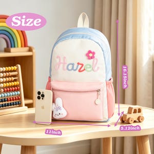 May include: A light blue, white, and pink backpack featuring the embroidered name Hazel, a small pink flower, and a white plush rabbit charm. The backpack measures 41.02 cm in height, 27.94 cm in width, and 13 cm in depth.