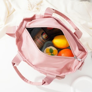 May include: A pink tote bag with a zipper closure, filled with food items. Inside the bag are a lemon, an orange, and a container with a sandwich. The bag has two handles and is sitting on a white surface.