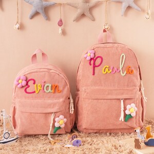 May include: Two pink corduroy backpacks, one smaller and one larger, with the names "Evian" and "Paula" embroidered in colorful yarn. Each backpack features a flower applique and a front pocket. The backpacks are designed for children.