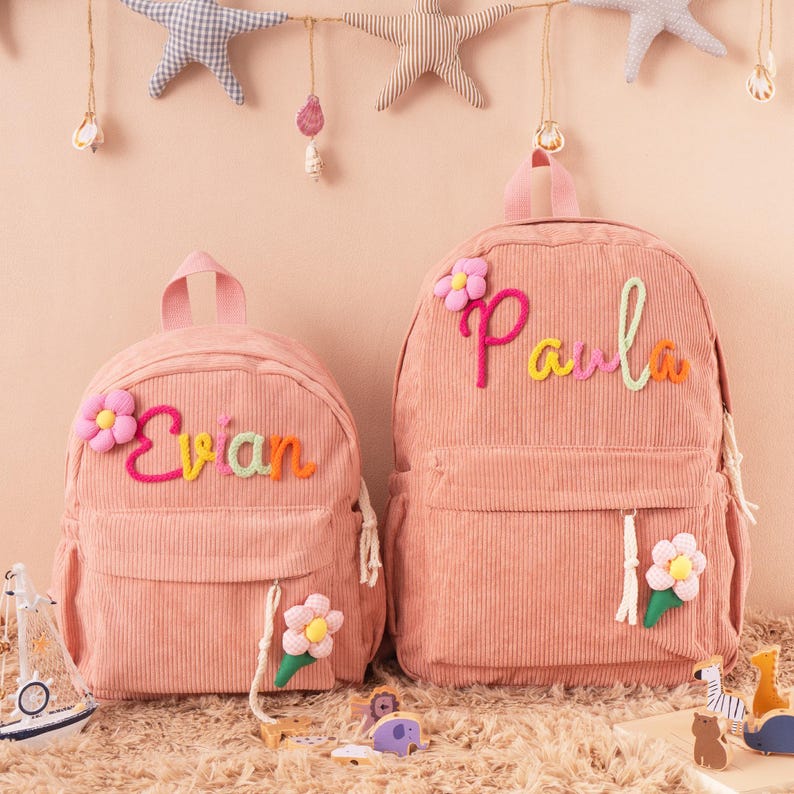 May include: Two pink corduroy backpacks, one smaller and one larger, with the names "Evian" and "Paula" embroidered in colorful yarn. Each backpack features a flower applique and a front pocket. The backpacks are designed for children.