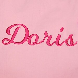May include: A close-up of a pink fabric with the name "Doris" embroidered in a vibrant, cursive script. The embroidery is a darker shade of pink, creating a contrast against the lighter pink background.