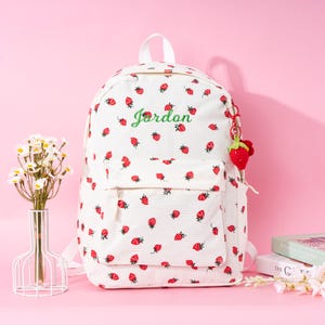 May include: White canvas backpack with a red strawberry pattern and the name "Jordan" embroidered in green. The backpack has a small front pocket and a strawberry keychain.